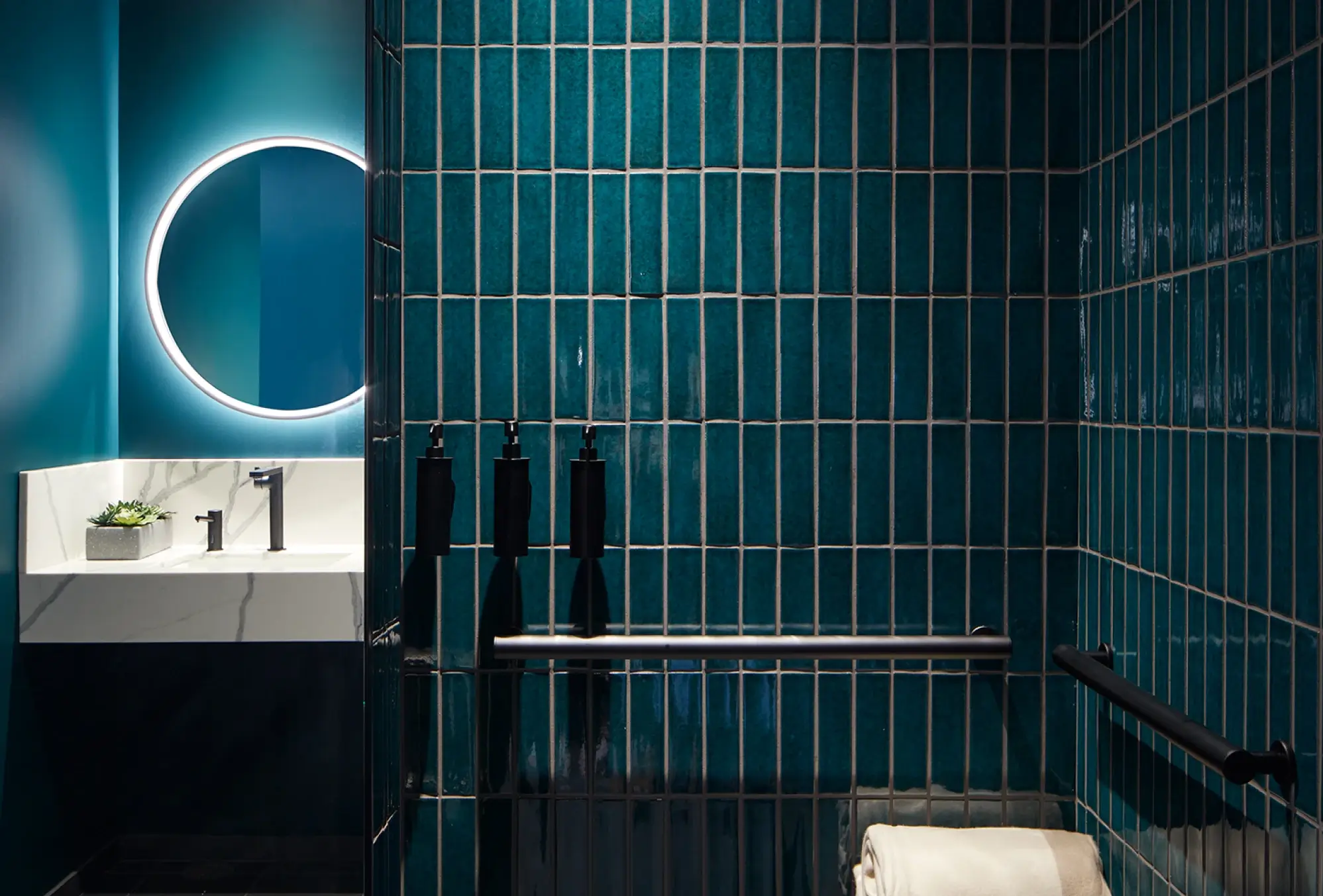 Stylish office bathroom with glowing mirror, teal vertical tiles, and matte black soap dispensers