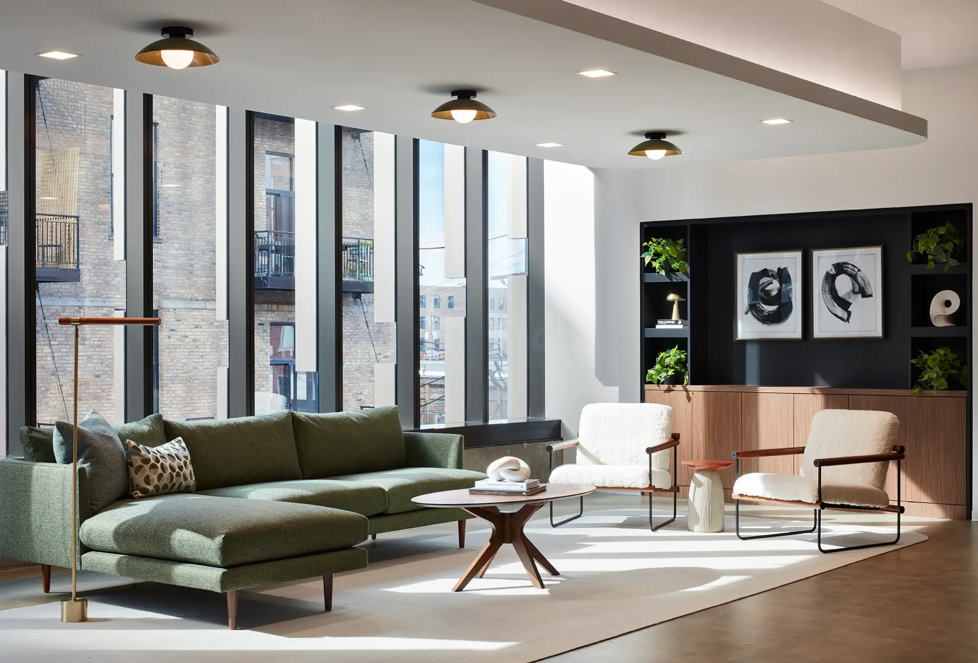 Daylit lounge with green sectional sofa, sculptural chairs, and floor-to-ceiling windows facing brick building
