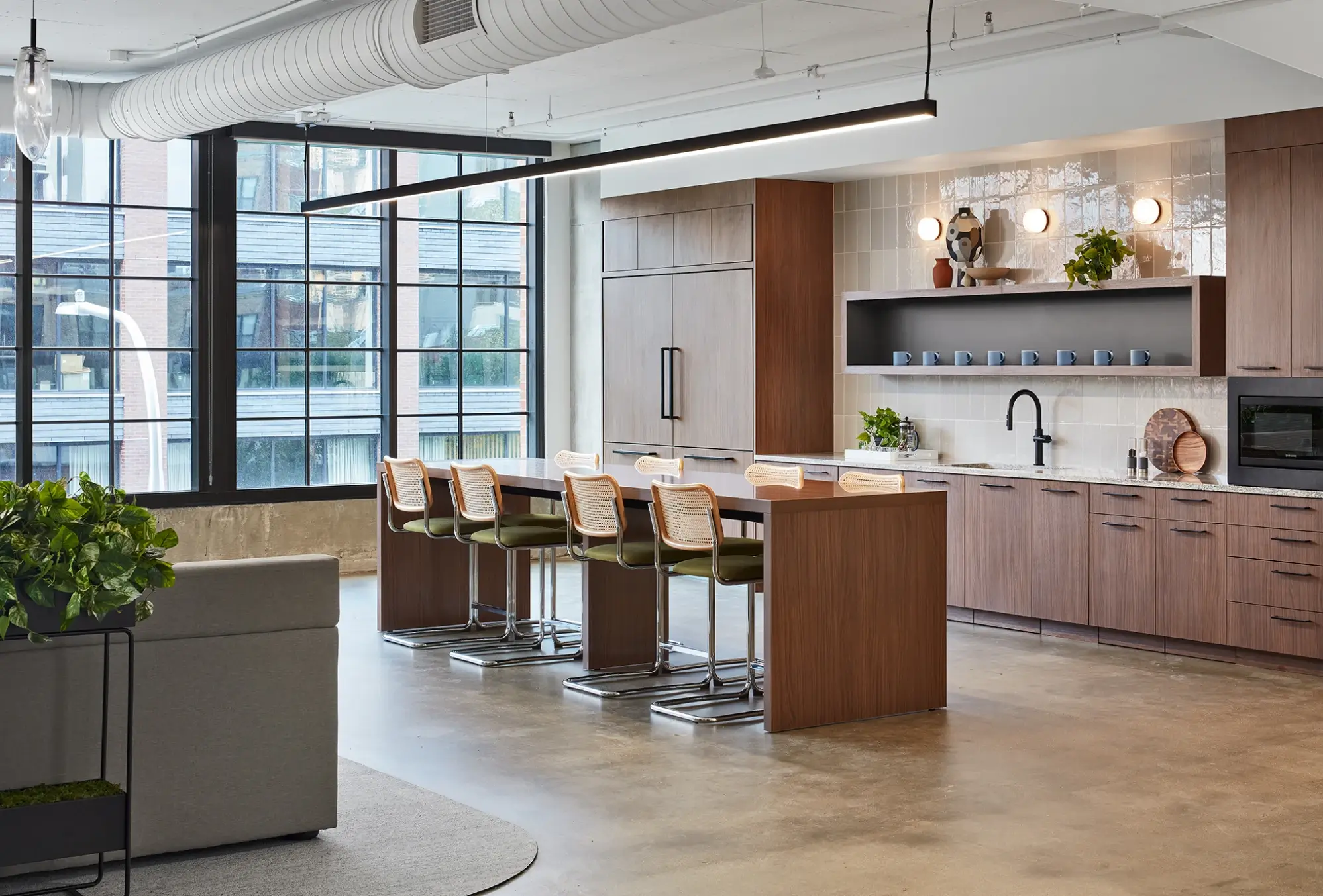 Office kitchen with wood cabinetry, bar-height island, green chairs, and industrial window grid backdrop