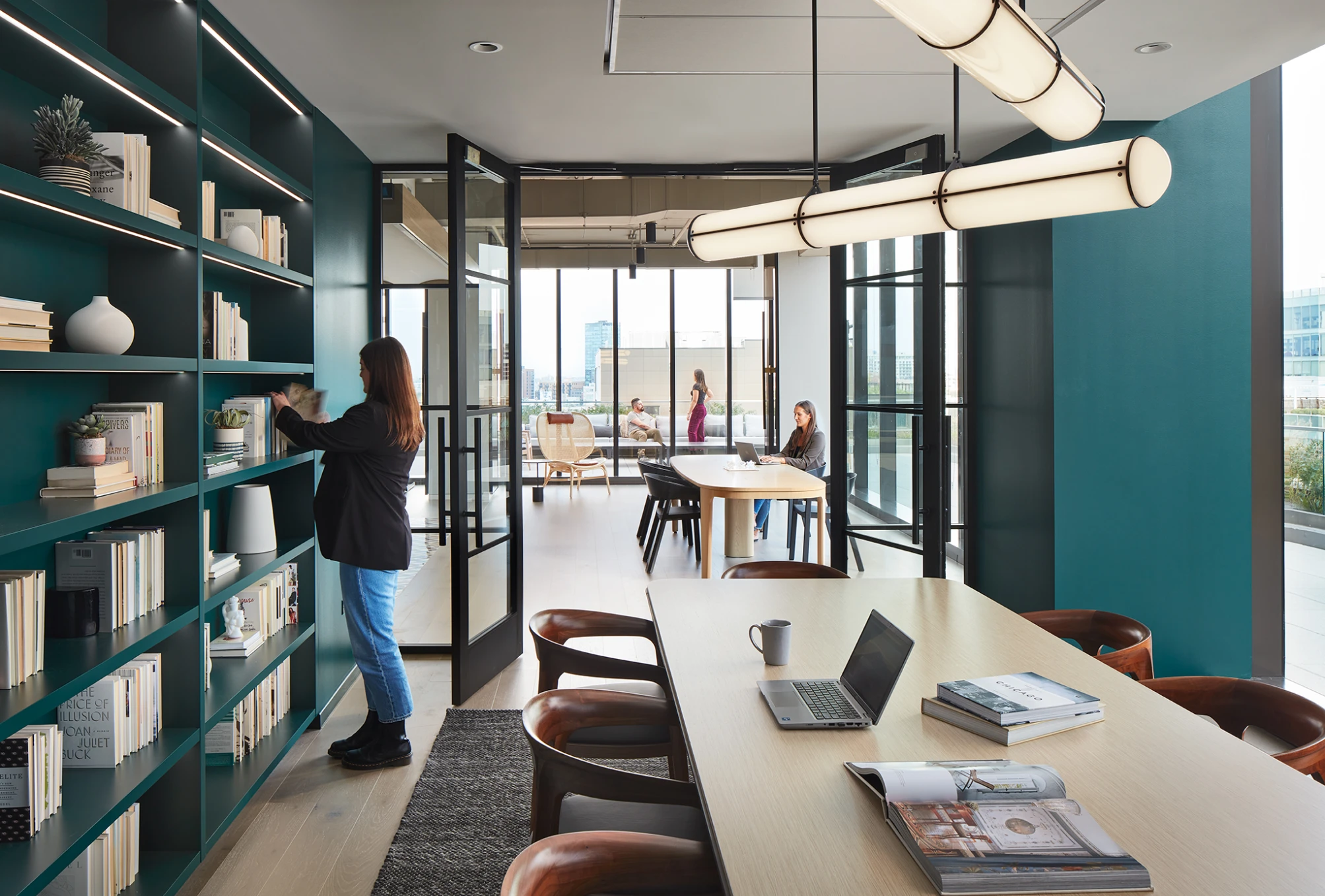Modern office lounge with teal bookshelves, glass doors, and people working in natural light